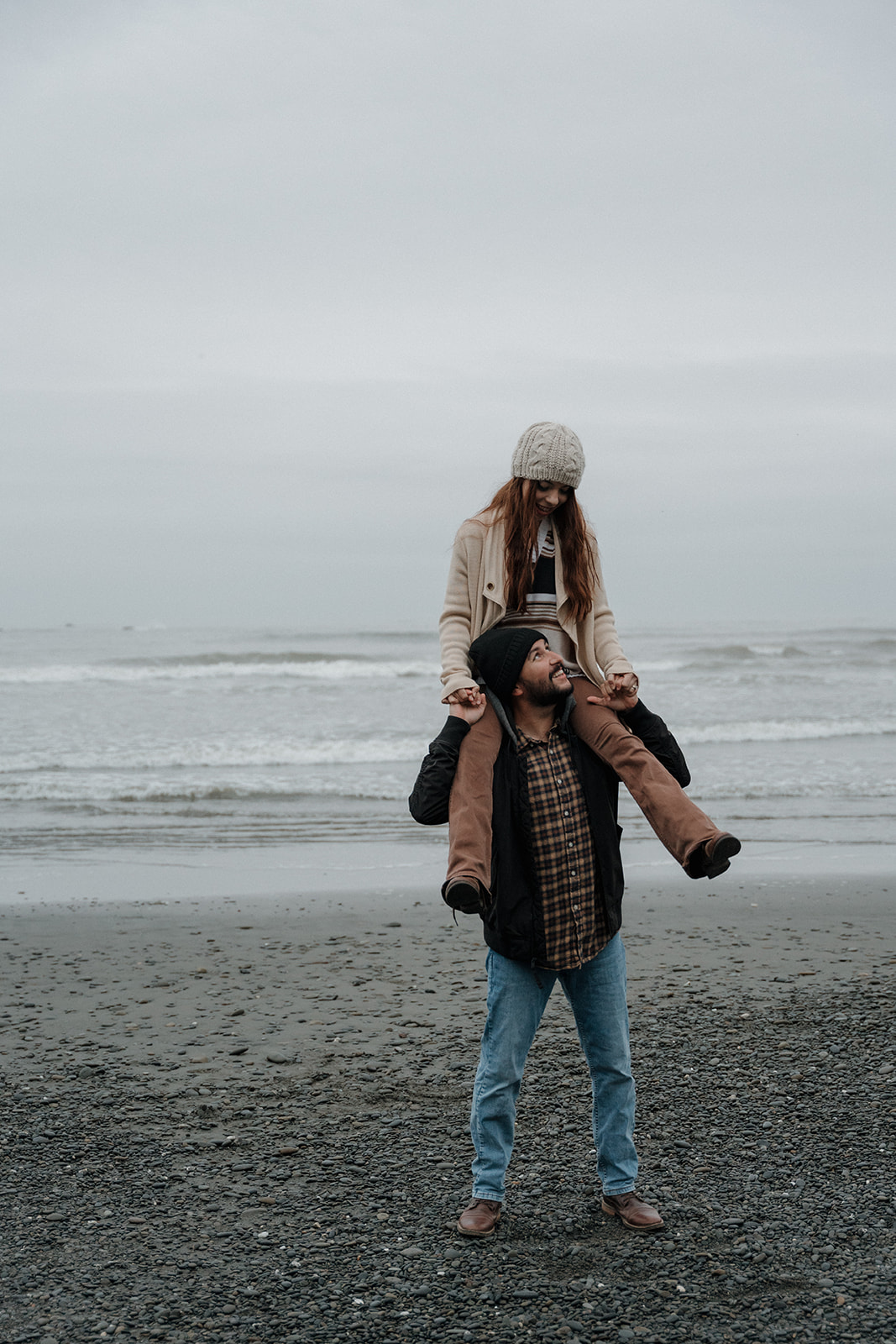 A man and a woman in winter clothing hold hands while standing on a rocky beach with waves and a cloudy sky in the background fir a washington engagement