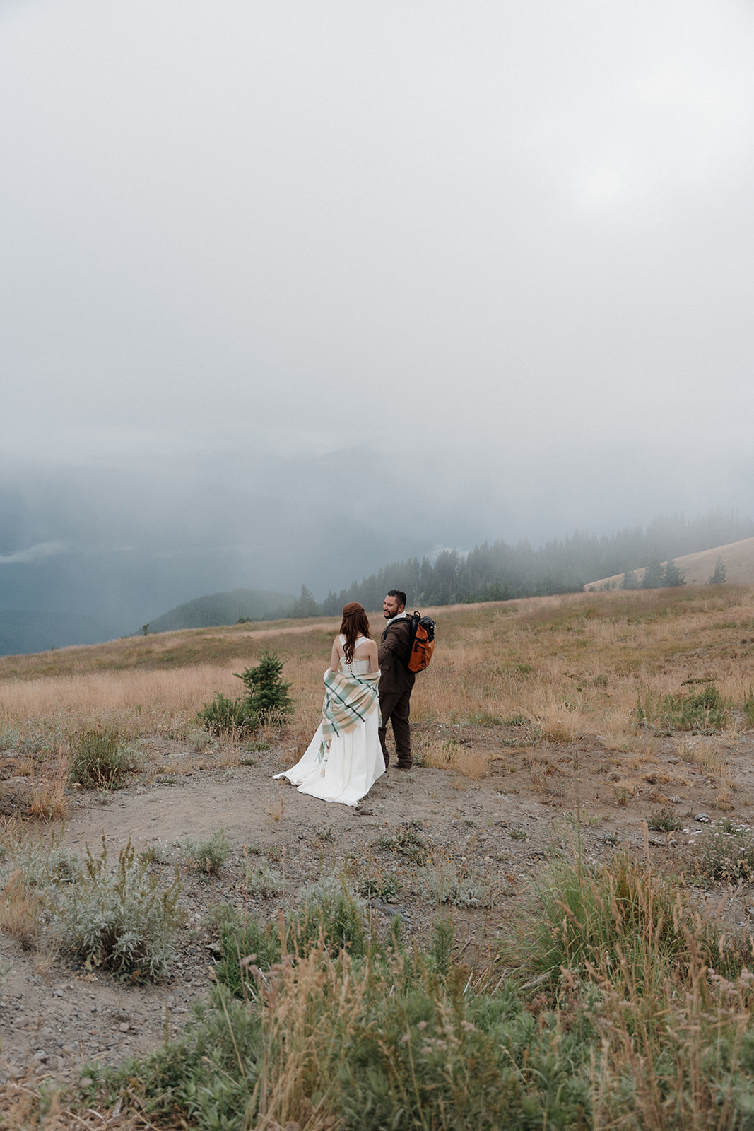 A bride in a white dress and a groom with a backpack stand on a grassy hillside under a foggy sky for their PNW fall elopement