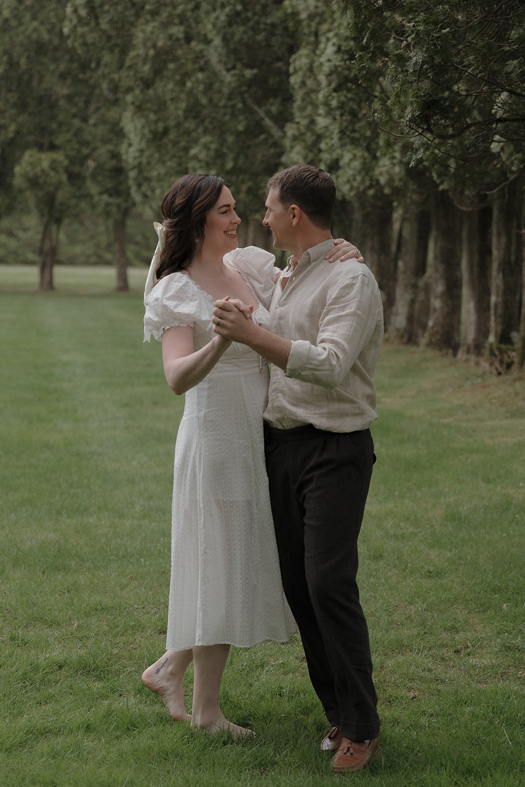 A couple dances barefoot on grass near a row of trees, smiling at each other; the woman wears a white dress, and the man wears a light shirt and dark pants for their seattle engagement photos
