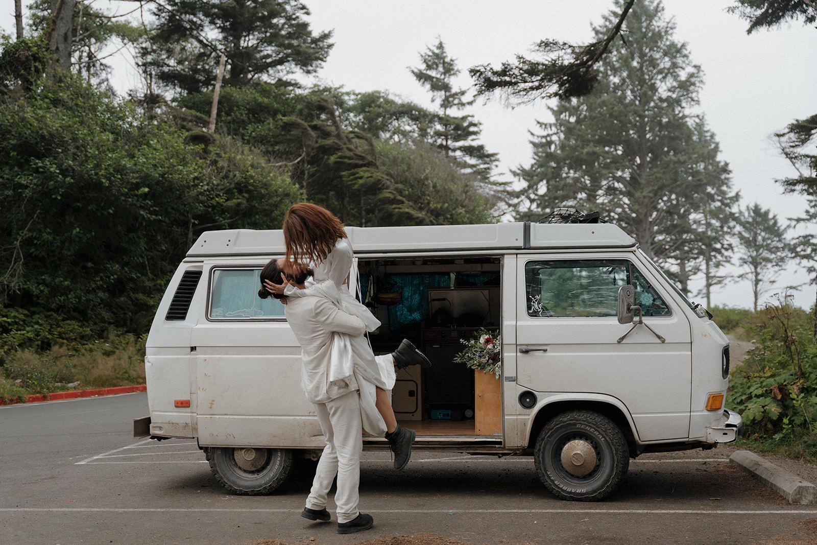 A person in a white outfit lifts another person in front of a white van parked near trees, with the van door open revealing items inside.