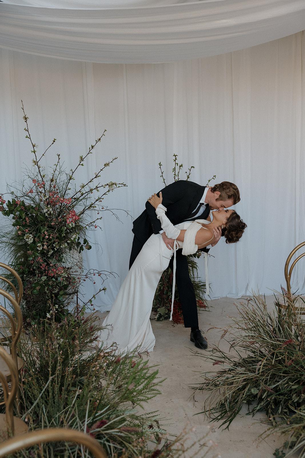 A bride and groom walk down an aisle decorated with greenery and flowers under white drapes, holding hands and raising their arms in celebration.